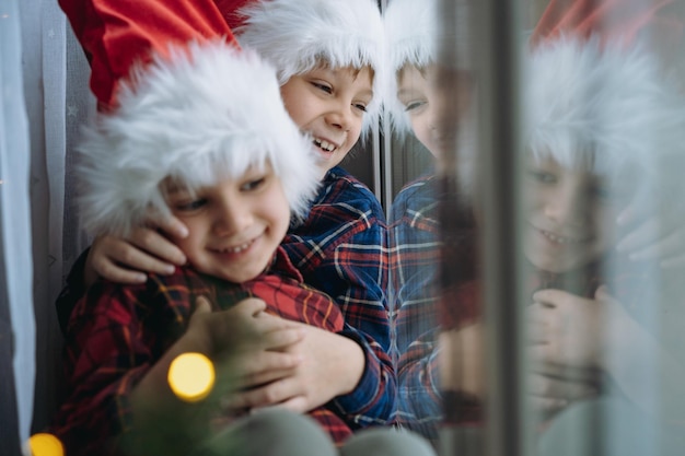 Enfants portant des chapeaux snta et des chemises à carreaux assis serrant près de la fenêtre attendant Noël