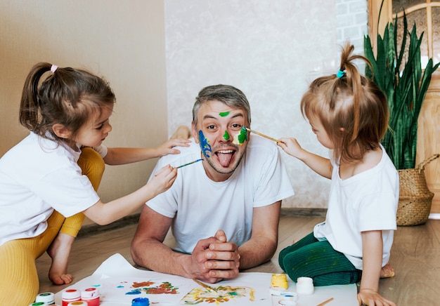 Les enfants s'amusent avec leur père, les filles dessinent sur la peau du visage d'un homme avec des peintures colorées, la créativité et l'imagination de la fête des pères et le concept de la famille