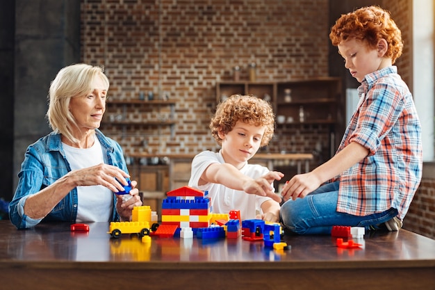 Les enfants sont l'arc-en-ciel de la vie. Grand-mère consciente regardant ses petits-enfants assis à une table et jouant avec un ensemble de jouets en plastique colorés