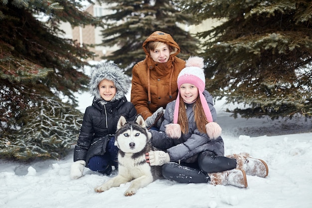 Les enfants sont assis dans la neige et caressent un chien husky. Les enfants sortent et jouent avec un chien husky en hiver. Promenade dans le parc