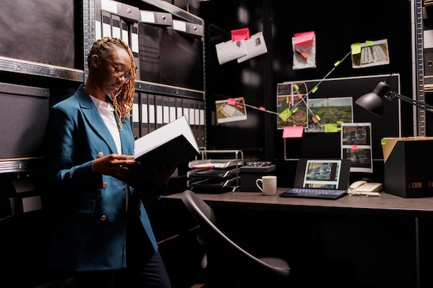 Une enquêteuse de police afro-américaine lit le dossier dans la salle de bureau. Détective privé tenant un dossier et analysant un rapport d'enquête près du tableau avec un schéma de connexion d'indices