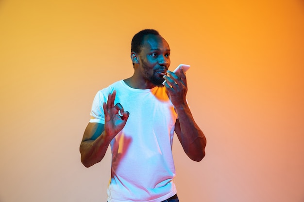 Enregistrement d'un message vocal. Portrait moderne de l'homme afro-américain sur fond de studio orange dégradé au néon. Beau modèle afro. Concept d'émotions humaines, d'expression faciale, de ventes, d'annonces.
