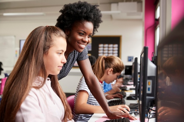 Enseignant aidant la ligne d'élèves de sexe féminin d'élèves du secondaire travaillant sur des écrans dans la classe d'informatique