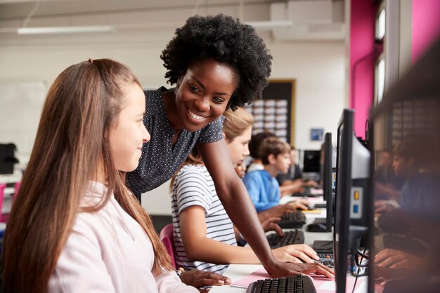 Enseignant aidant la ligne d'élèves de sexe féminin d'élèves du secondaire travaillant sur des écrans dans la classe d'informatique