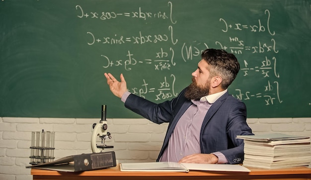 Enseignant hipster charismatique s'asseoir à table fond de tableau de classe Conversation éducative Parler aux étudiants ou aux élèves Concept d'enseignant d'école Un enseignant barbu raconte une histoire intéressante