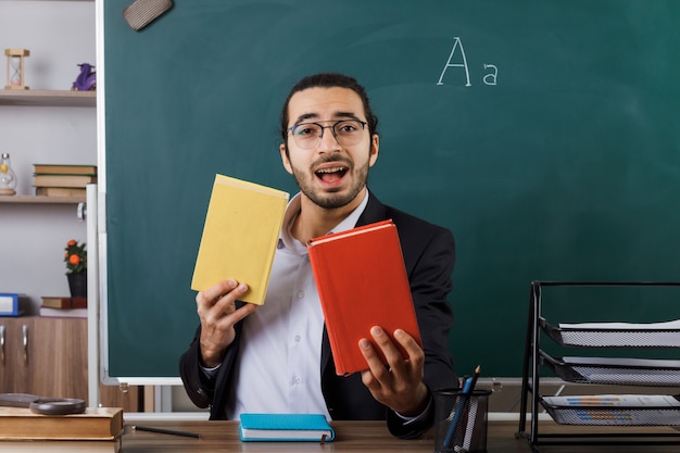 Enseignant joyeux portant des lunettes tenant un livre assis à table avec des outils scolaires en classe