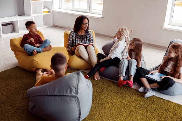 Enseignant à lunettes assis sur un pouf et lisant pour les écoliers