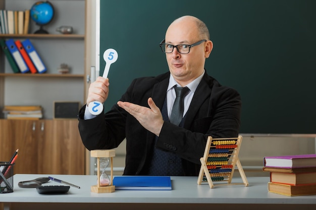 Un enseignant portant des lunettes assis au bureau de l'école avec un registre de classe devant le tableau noir dans la salle de classe tenant des plaques d'immatriculation expliquant la leçon heureuse et positive