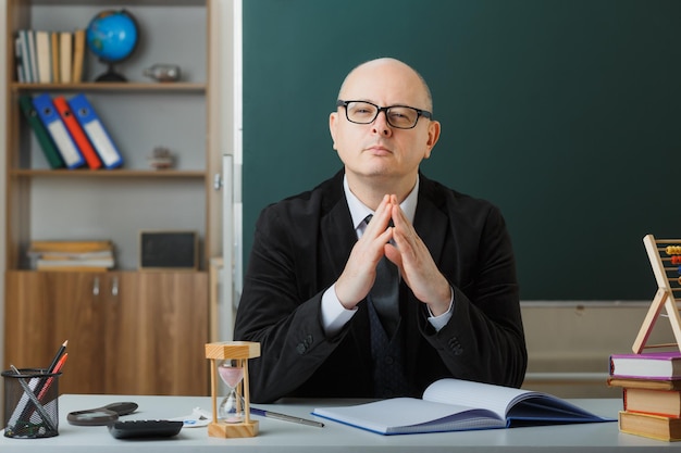 Enseignant portant des lunettes vérifiant le registre de classe regardant la caméra tenant les paumes ensemble positif et apaisé assis au bureau de l'école devant le tableau noir dans la salle de classe