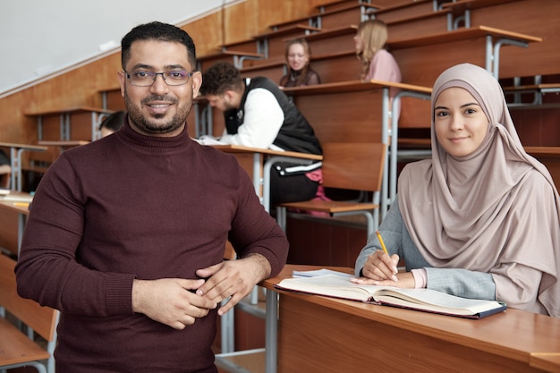 Enseignant de sexe masculin réussi debout près d'un bureau où une jeune étudiante en hijab est assise et prend des notes en lisant un livre pendant la leçon