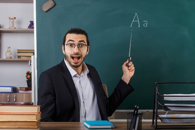 Un enseignant surpris portant des lunettes pointe sur un tableau noir avec un bâton de pointeur assis à table avec des outils scolaires en classe