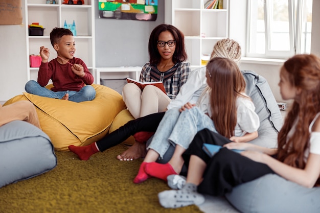 Enseignante assise sur un pouf et lisant pour les écoliers