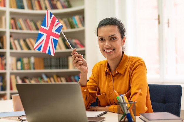 Enseignante positive avec le drapeau de la grande-bretagne travaillant avec un ordinateur portable à l'intérieur de la bibliothèque