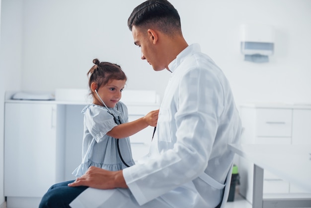 Enseigne à la fille comment utiliser le stéthoscope. Un jeune pédiatre travaille avec une petite visiteuse à la clinique.