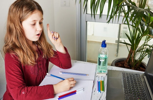 Enseignement à distance en ligne. Écolière étudie à la maison avec un ordinateur portable et fait ses devoirs. Livres d'entraînement et feutres colorés sur la table, gel avec de l'alcool à 70 pour cent