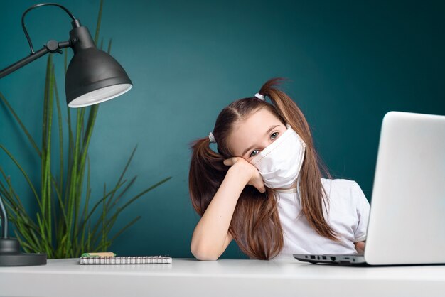 Enseignement à Distance En Ligne. écolière En Masque Médical étudie à La Maison, Travaille Sur Un Ordinateur Portable Et Fait Ses Devoirs. Concept De Quarantaine De Coronavirus