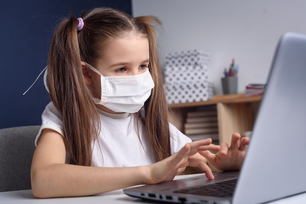 Enseignement à Distance En Ligne. écolière En Masque Médical étudie à La Maison, Travaille Sur Un Ordinateur Portable Et Fait Ses Devoirs. Concept De Quarantaine Covid