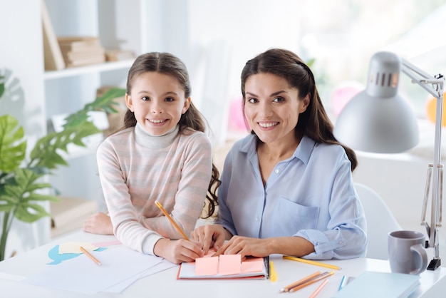 Enseignement à domicile. Belle mère et fille ravie joyeuse souriant et vous regardant tout en étudiant ensemble