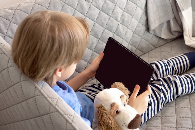 Enseignement et travail en ligne à distance. L'enfant étudie à distance depuis la maison sur le canapé. Les mains du garçon tiennent la tablette.