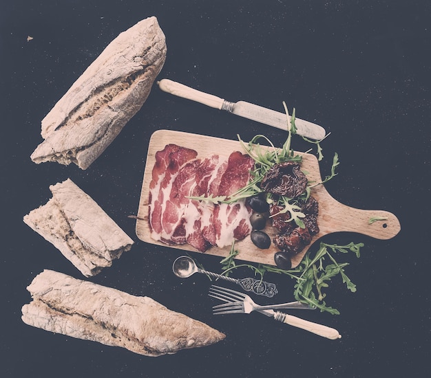 Photo ensemble d'apéritifs au vin vaisselle vintage baguette française brisée en morceaux tomates séchées olives viande fumée et roquette sur planche de bois rustique sur fond sombre