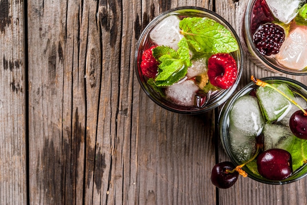 Ensemble de boissons rafraîchissantes traditionnelles d'été mojito aux baies. Au citron vert, feuilles de menthe, mûres, framboises et cerises. Avec les ingrédients sur une vieille table en pierre rustique en bois. vue de dessus