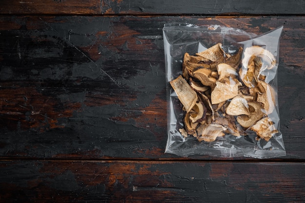 Ensemble de champignons sauvages séchés, sur le vieux fond de table en bois sombre, dans un emballage en plastique, vue de dessus à plat, avec espace pour fond de texte