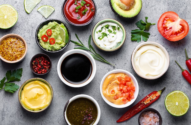 Ensemble de différentes sauces dans des bols et ingrédients sur fond de béton rustique gris, vue de dessus. Tomate ketchup, mayonnaise, guacamole, moutarde, sauce soja, pesto, sauce fromage - assortiment de trempettes