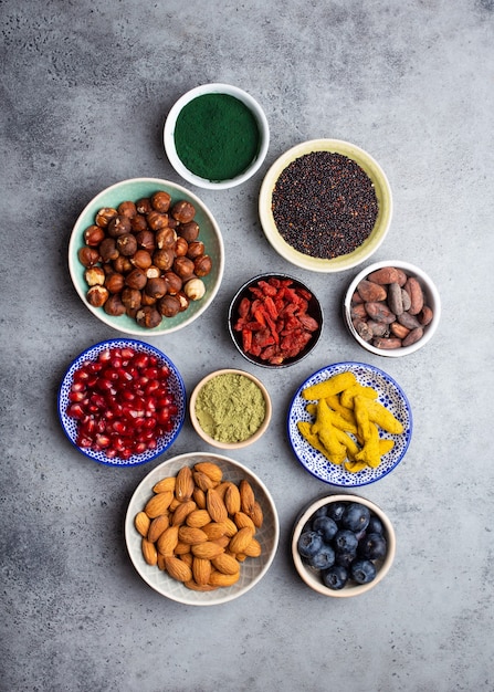 Ensemble de différents superaliments dans des bols sur fond gris pierre : spiruline, baie de goji, cacao, poudre de thé vert matcha, quinoa, graines de chia, myrtilles, noix pour une vie saine et heureuse, vue de dessus, gros plan