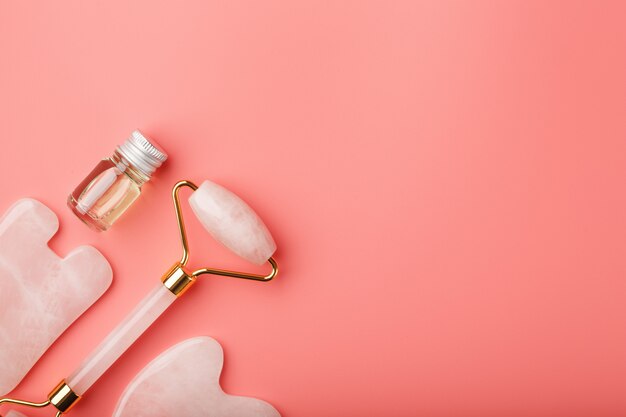 Un ensemble d'outils pour la technique de massage du visage Gua Sha en quartz rose naturel sur fond rose. Roller, pierre de jade et huile dans un bocal en verre pour les soins du visage et du corps.