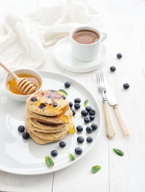 Ensemble de petit-déjeuner crêpes aux bleuets avec des baies fraîches feuilles de menthe miel et tasse de café sur fond de bois blanc