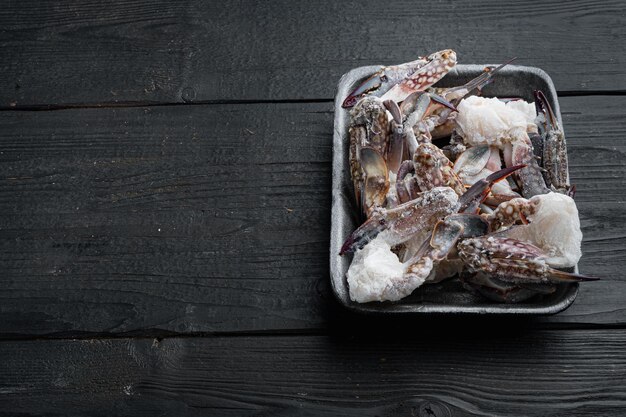 Ensemble de pièces de crabe de natation bleu brut congelé, dans un plateau en plastique, sur fond de table en bois noir, avec fond et espace pour le texte