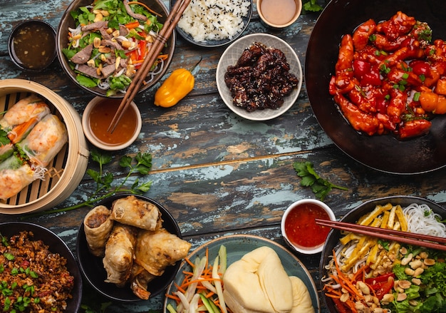 Ensemble de plats chinois assortis sur table : poulet aigre-doux dans un wok, dim sum, rouleaux de printemps, nouilles, salade, riz, petits pains cuits à la vapeur. Dîner ou buffet de style asiatique avec vue de dessus et espace pour le texte