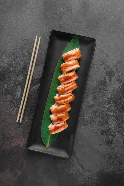 Ensemble de rouleaux de maki à l'anguille fumée et à la feuille de bambou verte dans une assiette en céramique noire avec des baguettes sur une vue de dessus de fond texturé gris foncé