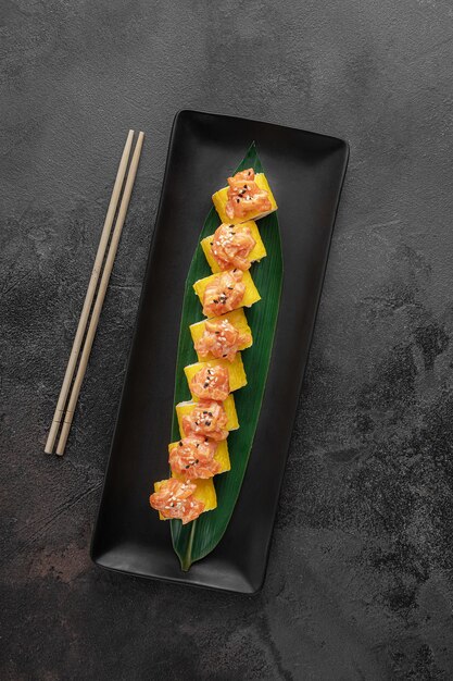 Ensemble de rouleaux de maki takuan jaunes avec du saumon à l'avocat de crevettes tigrées et une feuille de bambou verte dans une assiette en céramique noire avec des baguettes sur une vue de dessus de fond texturé gris foncé