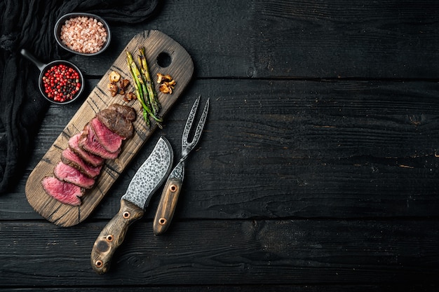 Ensemble de steaks de boeuf filet grillé, avec l'oignon et les asperges, sur une planche de service en bois, avec un couteau à viande et une fourchette, sur une table en bois noir, vue de dessus à plat