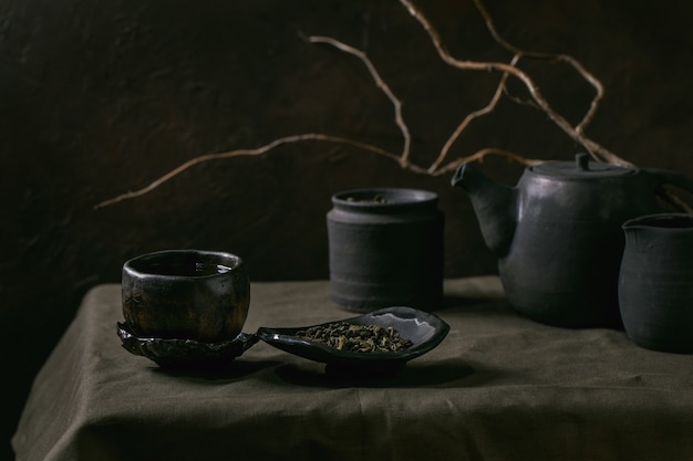 Ensemble de théière artisanale en céramique noire faite à la main, cruche, pot avec feuilles de thé, tasse wabi sabi, soucoupe pour la cérémonie du thé debout sur une nappe en lin avec branche sèche dans une pièce sombre.