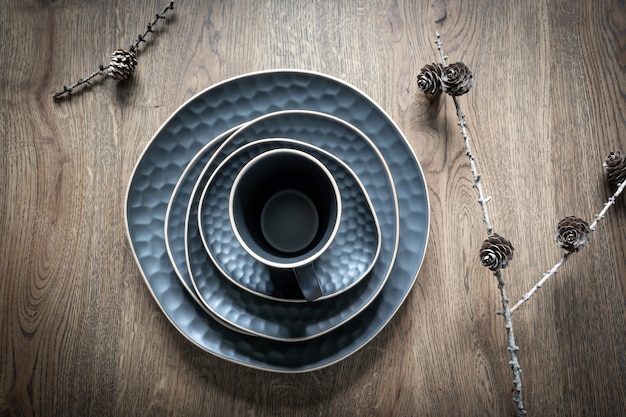 Ensemble de vaisselle bleu foncé : une tasse, une soucoupe, des assiettes, un bol et des cônes de mélèze secs sur des brindilles sur fond de bois. Décoration de table de Noël, vacances magiques