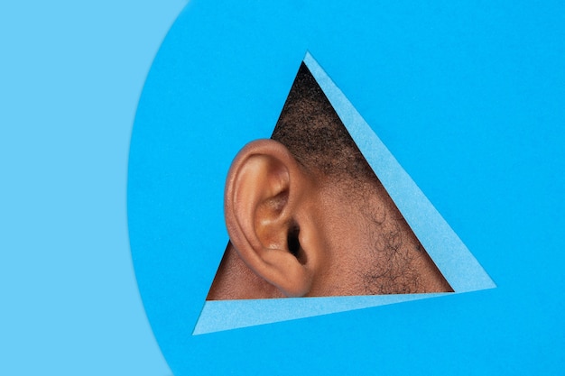 Photo entendre des secrets. l'oreille de l'homme afro-américain jette un coup d'œil à travers le triangle sur fond bleu. style géométrique à la mode, copyspace. couleurs vives. ventes, proposition, finances et affaires, concept de santé.