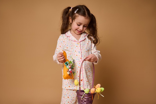 Enthousiaste adorable enfant Caucasien bébé fille en pyjama tenant un jouet de lapin et une décoration colorée d'œufs de Pâques sourit posant à la caméra isolé sur fond beige avec copie espace publicitaire