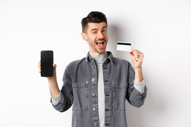 Enthousiaste beau mec montrant une bonne affaire, écran de smartphone vide et carte de crédit en plastique, vous faisant un clin d'œil et souriant, recommandant une offre, fond blanc.