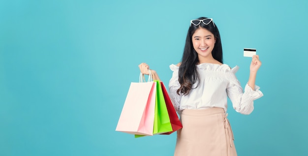 Enthousiaste belle femme asiatique tenant des sacs à provisions multicolores et carte de crédit sur bleu clair.