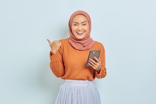 Enthousiaste belle femme musulmane asiatique en chandail marron et hijab tenant un smartphone et pointant le doigt sur l'espace de copie isolé sur fond blanc Concept de mode de vie musulman