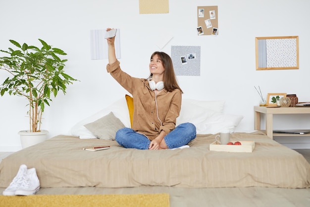 Enthousiaste belle jeune femme aux cheveux bruns assis avec les jambes croisées sur le lit et à l'aide de téléphone pour o