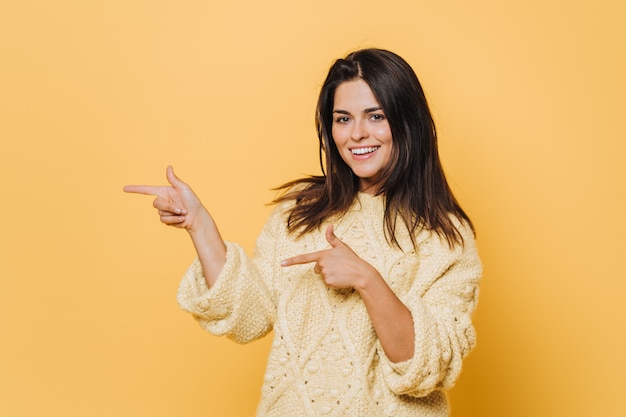 Enthousiaste belle jeune femme avec un large sourire, indique avec les deux doigts avant à l'espace vide, porte un pull jaune, isolé sur le mur jaune. Achetez cette chose à rabais ou en vente