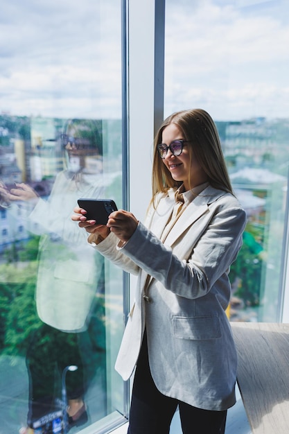 Enthousiaste belle jeune femme à lunettes parlant sur un téléphone portable et utilisant un ordinateur portable avec un sourire tout en étant assis sur son lieu de travail