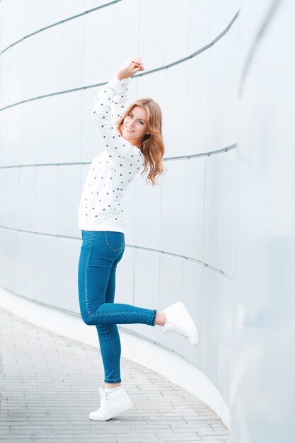 Enthousiaste belle jeune femme positive dans des vêtements à la mode s'amuser sur une chaude journée de printemps à l'intérieur près du mur moderne