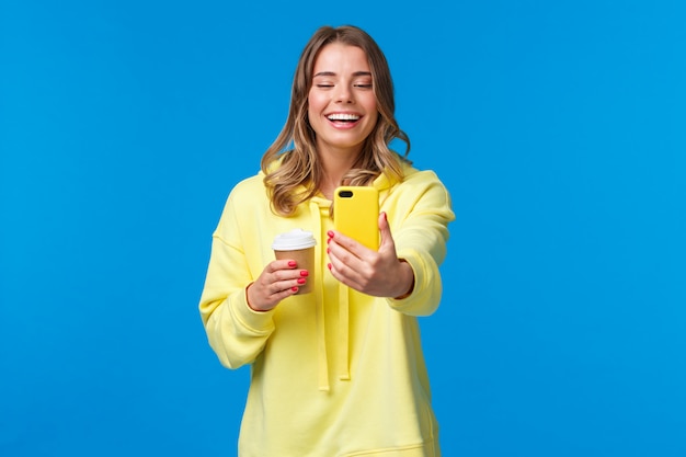 Enthousiaste blonde blonde caucasienne femme blogueuse enregistrer une vidéo ou prendre un selfie sur son téléphone, rire et sourire comme tenant une tasse de café à emporter, tenir le mur bleu