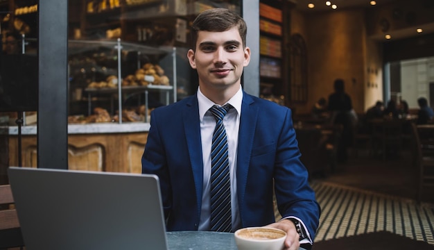 Enthousiaste directeur masculin brun vêtu d'un costume bleu et d'une cravate rayée avec une chemise blanche prenant une pause-café sur une terrasse de boulangerie confortable avec un ordinateur portable souriant et regardant la caméra