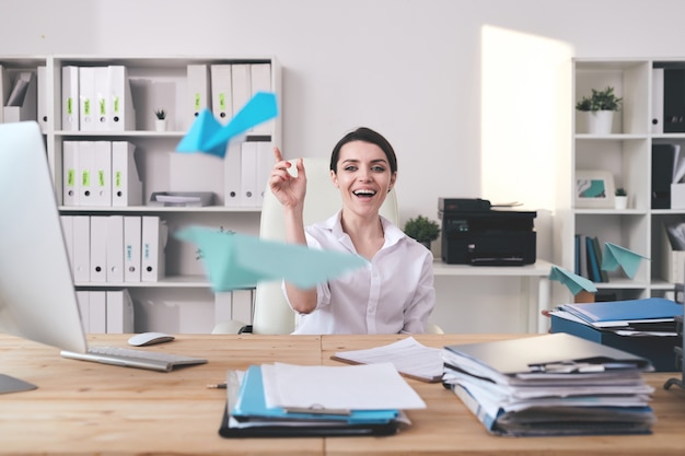 Enthousiaste excité jeune femme d'affaires assis au bureau plein de documents et jetant un avion en papier au bureau