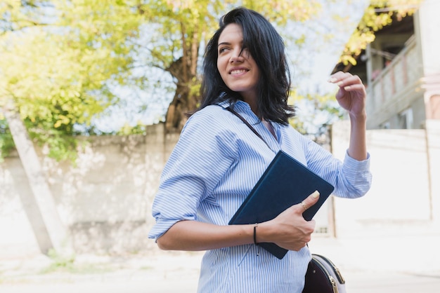 Enthousiaste femme d'affaires brune à l'aide d'un ordinateur portable pour planifier le temps de nouveaux projets en plein air Jeune étudiant intelligent portant une chemise bleue dans la rue